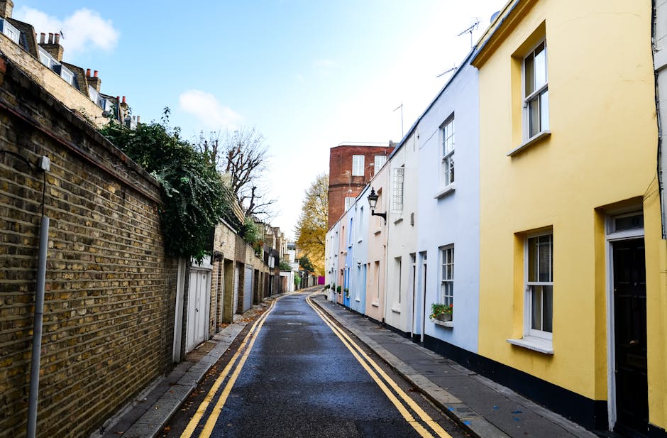 A narrow residential street with a slightly wet, dark asphalt surface marked with double yellow lines along the curb. On the left side, there is a brick wall topped with greenery and small garden sheds behind it. On the right side, there are rows of terraced houses with white and pastel yellow facades, each featuring sash windows, some with flower boxes. A black street lamp is mounted on one of the houses, and in the background, a multi-storey red brick building rises above the row of houses. The sky is partly cloudy with patches of blue visible, and the scene is illuminated by natural daylight. The setting suggests a quiet neighbourhood suitable for home relocation or furniture transport activities, with no moving equipment visible in this image itself, but it complements the context of house removals or packing and moving services provided by Haringey Man and Van.