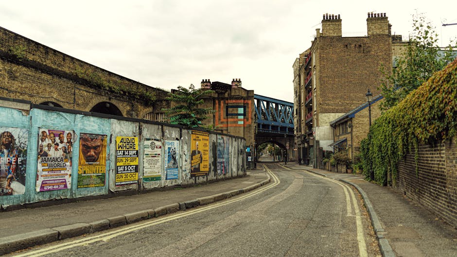 A narrow, curved residential street in Crouch End with double yellow lines along the edge of the pavement. On the left side, there is a long, weathered brick wall covered with colorful posters advertising events and music concerts, some of which are peeling. Behind the wall, parts of an older brick building and a large, multi-story structure with a dark roof are visible. To the right, a high brick wall with green foliage growing along its top runs adjacent to the sidewalk. An arched blue metal bridge spans over the street, connecting two buildings. Several multi-story brick buildings with small balconies and chimneys line the street, some featuring fire escapes. The scene appears during daylight with an overcast sky. This setting illustrates the tricky urban environment for home relocation and furniture transport, potentially requiring careful planning and maneuvering for removals, which Haringey Man and Van provides expertise for in such narrow street conditions.