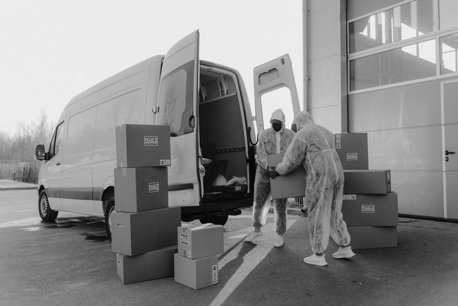 Two movers dressed in protective clothing, including white coveralls, masks, and hair covers, are seen loading cardboard boxes of various sizes into the back of a white panel van parked outside a modern building with large glass windows. The boxes, some of which are sealed with packing tape, are being carried carefully by the movers, who are positioned on a paved surface near the vehicle's open rear doors. Additional boxes are stacked on the pavement nearby, some resting on their sides and others upright. The scene reflects an organized loading process typical of home relocation or furniture transport services, with the exterior environment well-lit under natural daylight. The presence of packing materials and the careful handling of boxes highlight the logistical aspects of off-site packing and loading associated with professional removals, such as those offered by Haringey Man and Van, web address https://haringeymanandvan.com, as indicated in the page titled 'Haringey council loading bay permits for removal vans, HARINGEY.'