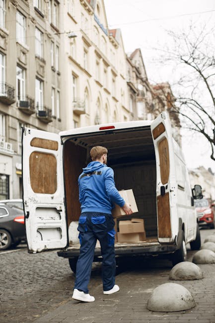 A man dressed in a blue jacket, dark blue trousers, and white sneakers is standing inside the open back of a white cargo van parked on a cobblestone street in Haringey. He is in the process of loading a cardboard box, which he is holding with both hands. Inside the van, there are additional cardboard boxes and packaging materials, including some wrapped in plastic. The van's interior has wooden panels on the sides and floor, and is surrounded by stone bollards on the pavement. The background features typical urban residential buildings with white facades, several windows, and balconies, under an overcast sky. The scene depicts a home relocation or furniture transport process, supported by the presence of packing materials and the loading activity, consistent with professional moving services by Haringey Man and Van.