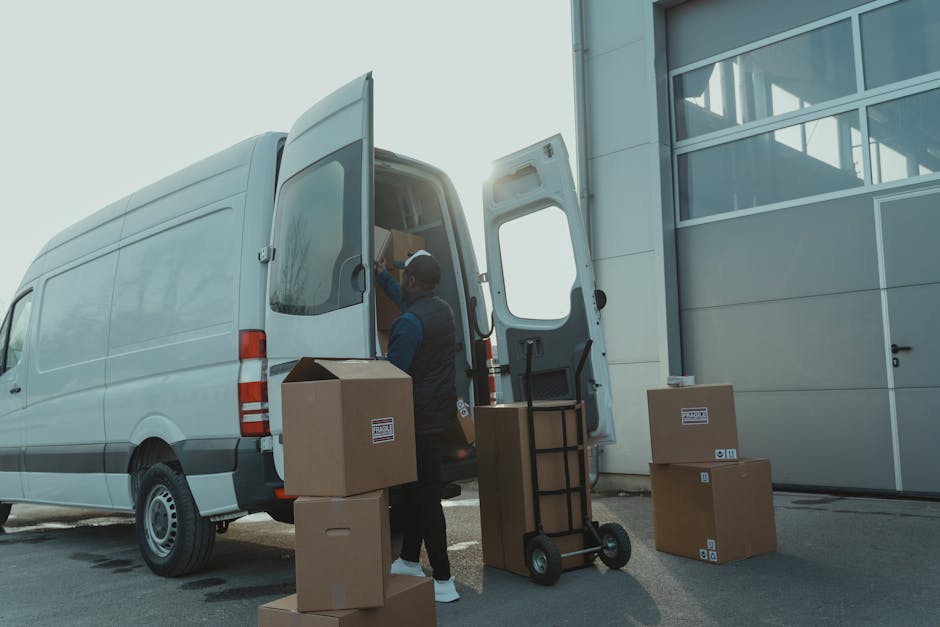 Two movers dressed in protective clothing, including white coveralls, masks, and hair covers, are seen loading cardboard boxes of various sizes into the back of a white panel van parked outside a modern building with large glass windows. The boxes, some of which are sealed with packing tape, are being carried carefully by the movers, who are positioned on a paved surface near the vehicle's open rear doors. Additional boxes are stacked on the pavement nearby, some resting on their sides and others upright. The scene reflects an organized loading process typical of home relocation or furniture transport services, with the exterior environment well-lit under natural daylight. The presence of packing materials and the careful handling of boxes highlight the logistical aspects of off-site packing and loading associated with professional removals, such as those offered by Haringey Man and Van, web address https://haringeymanandvan.com, as indicated in the page titled 'Haringey council loading bay permits for removal vans, HARINGEY.'