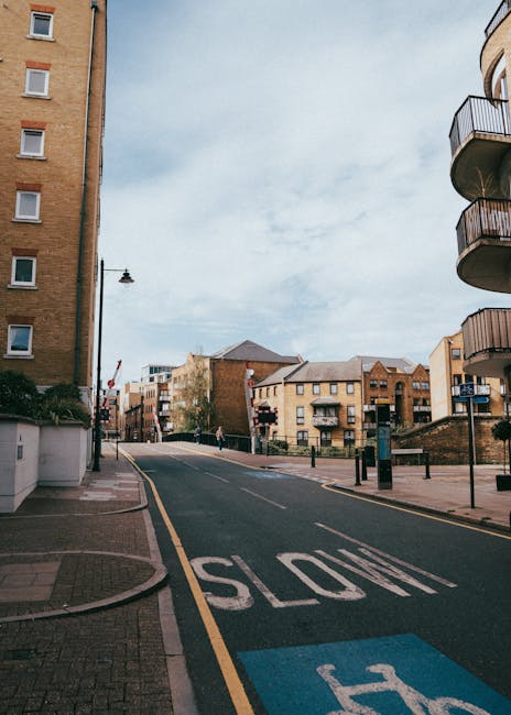 A residential street scene during daylight with a gently curving road marked with a large white 'STOP' sign and a blue accessible parking space. On the left, a tall multi-storey building with several small windows and a white boundary wall near the pavement. On the right, a modern apartment building features balconies with curved metal railings. In the background, a mix of brick and modern buildings with pitched roofs and trees lining the street are visible. The pavement is clean, and there are a few bollards lining the sidewalk. A tall streetlight stands along the road, and a few pedestrians are walking in the distance. This scene depicts an urban environment suitable for house removals and furniture transport, with clear access to and from the property, supporting efficient moving and packing services, as coordinated by Haringey Man and Van.