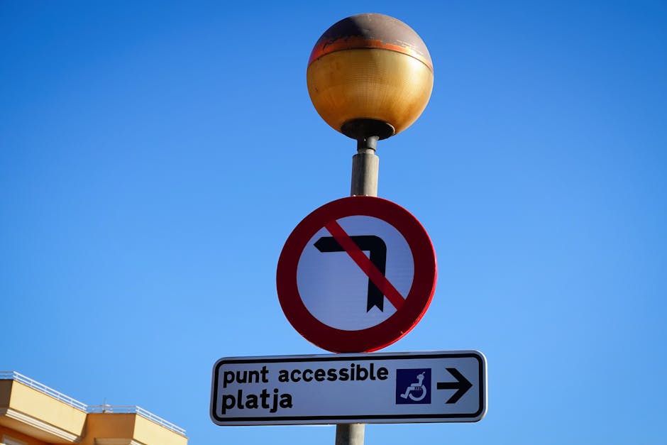 A street-facing lamppost with a rounded, multicoloured top, situated against a clear blue sky. Attached to the lamppost are multiple traffic and informational signs, including a red circle with a diagonal slash over a left turn arrow, indicating no left turns allowed. Below this, there is a rectangular sign with the words 'punt accessible platja' and an icon of a wheelchair, along with an arrow pointing to the right, guiding towards accessible parking or boarding points. The signs are prominently positioned at ground level, adjacent to a building with a beige facade visible in the background. This scene illustrates urban signage related to vehicle restrictions and accessible routes, which may be relevant to house removals or moving logistics in the area around Harringay Green Lanes, as referenced in the page title and website.