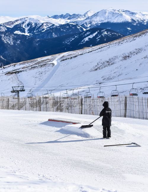 A person dressed in dark winter clothing and a helmet stands outdoors on a snow-covered slope, using a snow shovel to clear the snow around a ramp during a home relocation or skiing activity. In the background, there is a ski lift with empty chairs suspended above the snow, and a mountain range with snow-capped peaks under a clear sky. The scene is set on a bright, sunny day, with a wooden fence separating the slope from the ski lift area. This image captures the winter environment and the process of preparing a snowy outdoor space, which may relate to activities such as ski resort maintenance or winter sports facility management, consistent with the context of equipment and movement in snowy conditions.