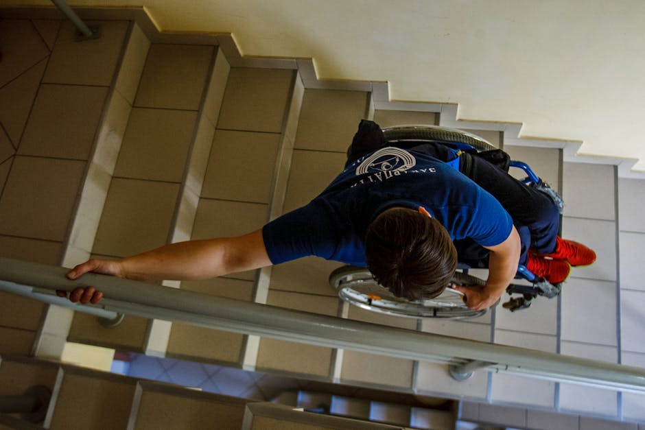 A person in a wheelchair, wearing a blue t-shirt and red socks, is carefully navigating down a staircase inside a building for a home relocation. The individual is holding onto a metal handrail with one hand while using the other to stabilize the wheelchair, which is positioned on each step. The stairs are made of beige tiles with white risers, and a beige wall is visible to the side. The wheelchair has a black backrest, and personal belongings are secured in a blue backpack attached to the back. The lighting is natural, coming from an unseen source, illuminating the staircase area. This scene depicts the logistical challenges of moving within high-rise buildings or multi-story homes, emphasizing the importance of professional moving and transport services, such as those provided by Haringey Man and Van, to manage stairs and lift difficulties during furniture transport and packing processes.