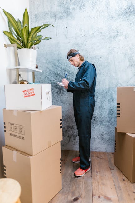 A young man and woman are seated on the wooden floor of a room, surrounded by packed cardboard boxes and household items, indicating a home relocation or packing process. The man has dark hair tied back and is wearing a black T-shirt and beige trousers, while the woman has curly blonde hair, is dressed in a white T-shirt with a pink and white jacket, and holds a notebook or clipboard, possibly taking notes related to the move. Behind them, there is a bed with white bedding, a closed green hard-shell suitcase resting on the bed, and a window with sheer curtains letting in natural light, illuminating the space. Several cardboard boxes are stacked near the bed, some taped shut, suggesting they are ready for transport or storage. The overall scene reflects the process of packing and preparing for a house move, with a focus on the moving logistics managed by Haringey Man and Van, a professional removal service specializing in furniture transport and home relocation within the Tottenham area.