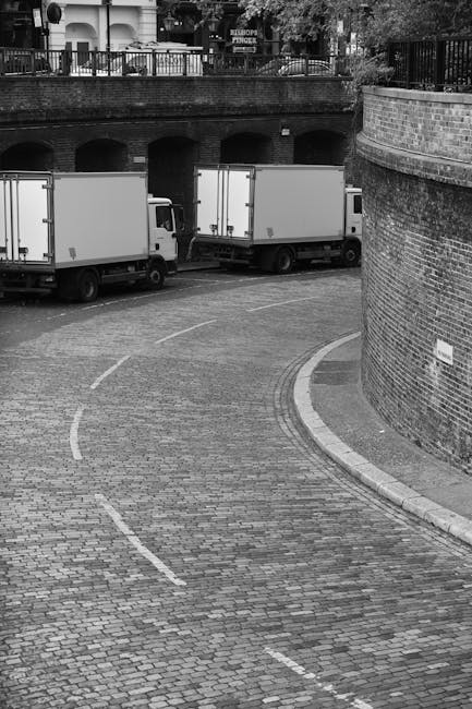 A black-and-white photograph of a narrow, curved cobblestone street in an urban area, with a brick retaining wall on the right side and a metal fence above it. In the background, two large white box trucks are parked parallel to each other on the street, with the rear doors facing toward a stone-arched structure. Inside the open doors of the trucks, visible are partially loaded furniture items, including cardboard boxes, wrapped chairs, and small household appliances. The trucks are positioned to facilitate a home relocation or furniture transport process, likely coordinated by Haringey Man and Van for same-day removals in Haringey. The scene is illuminated by natural daylight, and the environment suggests an active loading or unloading operation consistent with professional moving services amid urban infrastructure.