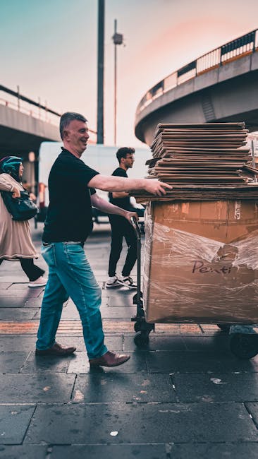 A man in a black shirt and blue jeans is pushing a large moving trolley loaded with multiple stacked cardboard boxes and wrapped furniture, situated outdoors under an overpass with a curved bridge structure in the background. Several other people are visible nearby, some walking past, indicating a busy urban environment. The trolley is positioned on a paved surface, and the scene is captured during daylight with natural lighting. This image reflects aspects of home relocation and furniture transport, highlighting the loading process involved in house removals, as operated by Haringey Man and Van, consistent with their services in same day removals to avoid delays and hidden fees.