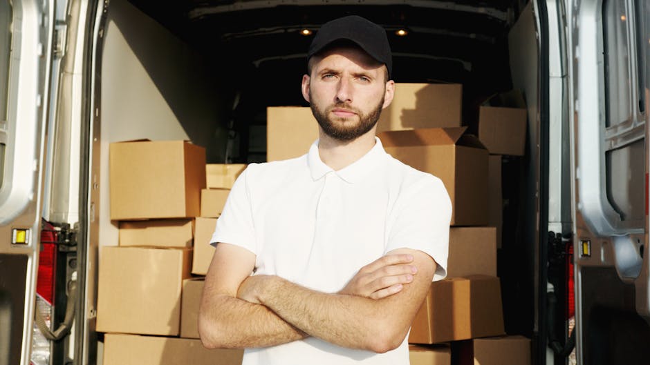 A young man with a serious expression, wearing a white polo shirt and a black cap, stands inside the back of a large moving van filled with numerous cardboard boxes of various sizes, some wrapped in p