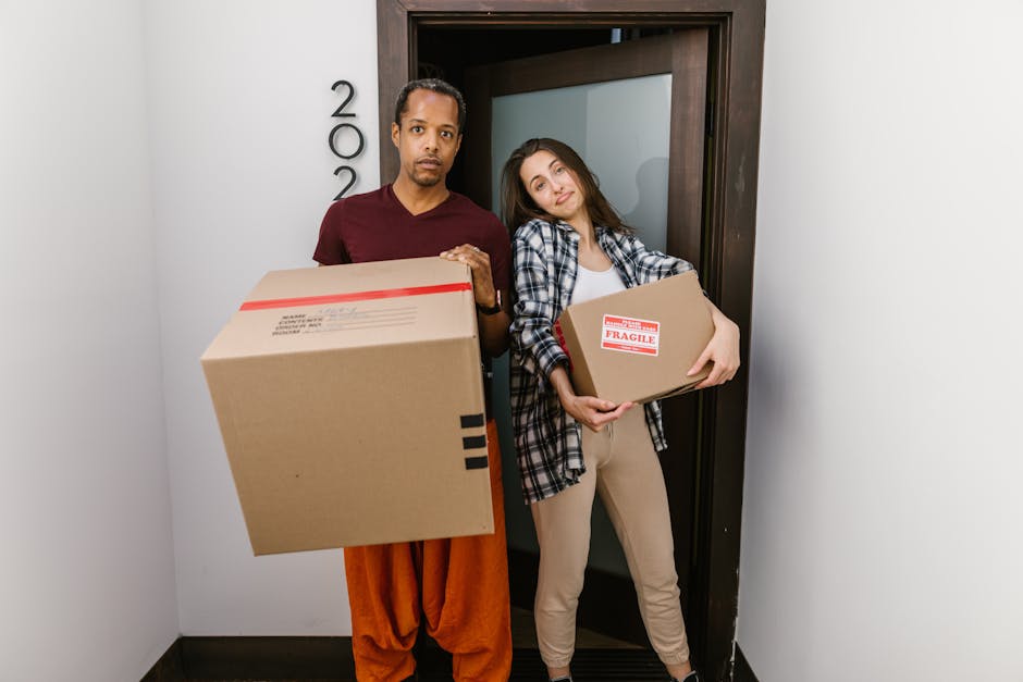 Close-up of a person holding an open cardboard box labeled 'FRAGILE' with red and white handling instructions, in a room filled with stacked cardboard boxes during a house removal or moving process. T