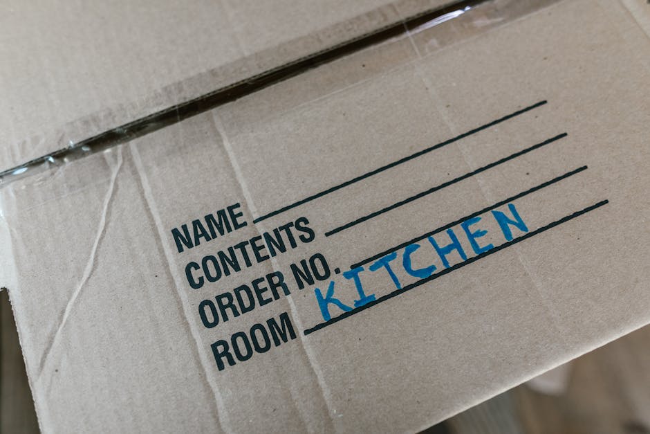 Close-up view of a cardboard moving box with printed sections for label details including 'NAME', 'CONTENTS', 'ORDER NO.', and 'ROOM', with 'KITCHEN' handwritten in blue marker next to the 'ROOM' label. The box is sealed with clear packing tape and positioned outdoors on a paved surface, possibly in a driveway or loading area. In the background, part of a moving van or vehicle used for home relocation is visible, indicating the process of furniture transport and packing during a house move. The scene reflects a professional removals service, such as Haringey Man and Van, involved in loading packed boxes into a van as part of a household move or home relocation task.