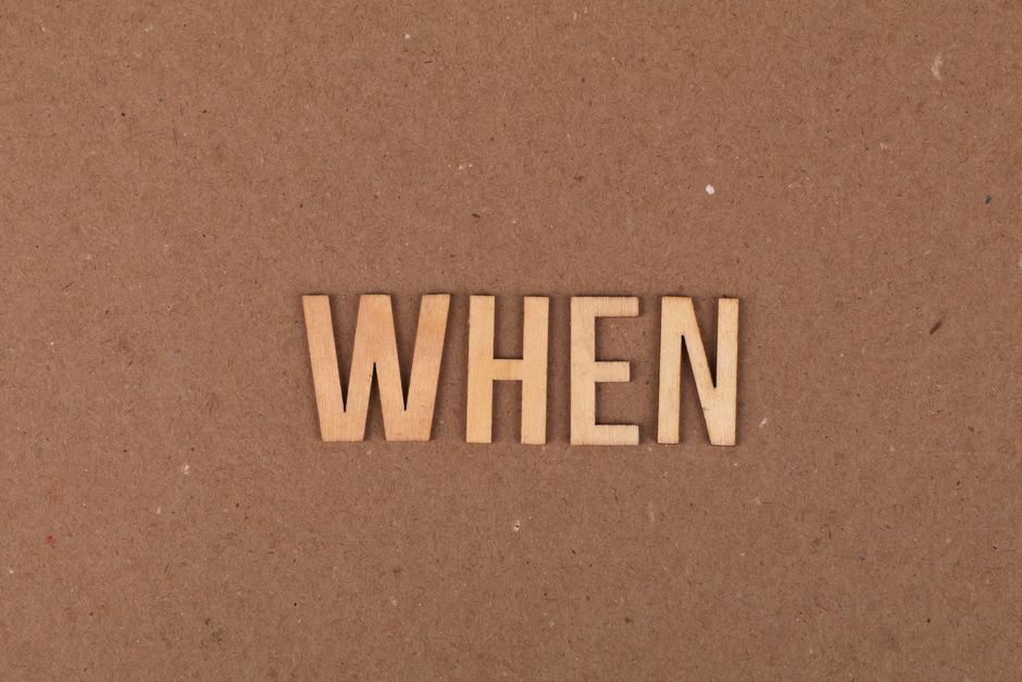 Close-up image of the word 'WHEN' spelled out with light wooden letter tiles arranged on a plain, brown cardboard surface. The background appears smooth with subtle texture, and the lighting is even, highlighting the natural wood finish of the tiles. The letters are centered in the image, creating a clear focus on the word. This visual can relate to topics such as timing, scheduling, or planning in home relocation or moving services, as shown on the Haringey Man and Van website, particularly in the context of handling booking issues or cancellations during moves involving packing, loading, and furniture transport.