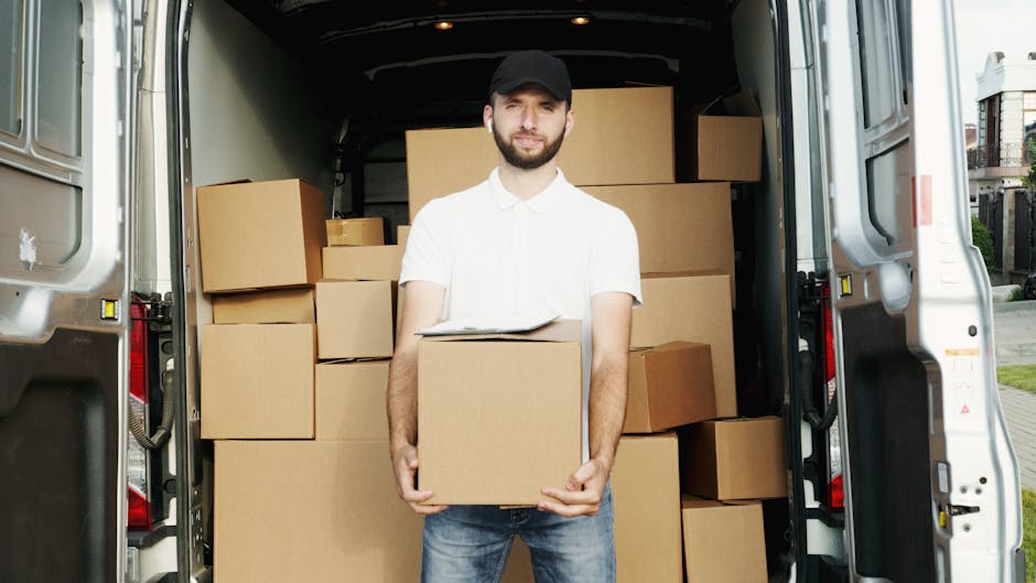 A man with a beard wearing a black cap, white polo shirt, and jeans stands inside the open rear of a moving van, holding a medium-sized cardboard box. The van's interior is filled with various cardboard boxes of different sizes, some stacked on top of each other, ready for a house removal or relocation. The van is parked outdoors on a paved driveway, with a residential area visible in the background, including trees and houses. The scene depicts a loading process typical of furniture transport and packing during home moves, supported by the presence of packing materials and the professional service provided by Haringey Man and Van, specializing in removals and house relocation support.