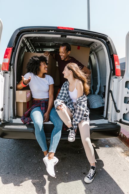 Three individuals are sitting at the back of a moving van, which is parked on the street near a sidewalk. The van's interior shows several cardboard boxes, some wrapped in packing paper and plastic, along with a few pieces of furniture or household items. One person is wearing a dark jacket, another is in a white top and light skirt, and the third has a plaid shirt tied around their waist, all engaged in conversation and smiling. The scene is outdoors with clear daylight, and the backdrop includes a pale blue sky. This image visually represents aspects of home relocation, packing, and furniture transport, illustrating the process of loading or unloading during a move, similar to services offered by Haringey Man and Van for house removals and house moves in the Alexandra Palace area.