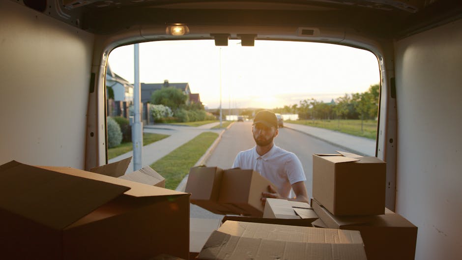 A man with a beard wearing a black cap, white polo shirt, and jeans stands inside the open rear of a moving van, holding a medium-sized cardboard box. The van's interior is filled with various cardboard boxes of different sizes, some stacked on top of each other, ready for a house removal or relocation. The van is parked outdoors on a paved driveway, with a residential area visible in the background, including trees and houses. The scene depicts a loading process typical of furniture transport and packing during home moves, supported by the presence of packing materials and the professional service provided by Haringey Man and Van, specializing in removals and house relocation support.
